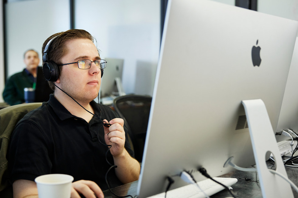 Exceptional Minds student Matthew Rohde is shown working at a Mac in a classroom on campus. Rohde is wearing a black polo and a pair of headphones.