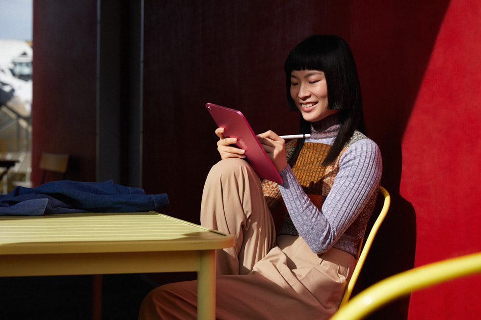 ผู้หญิงคนหนึ่งนั่งที่โต๊ะและใช้ Apple Pencil บน iPad สีชมพู