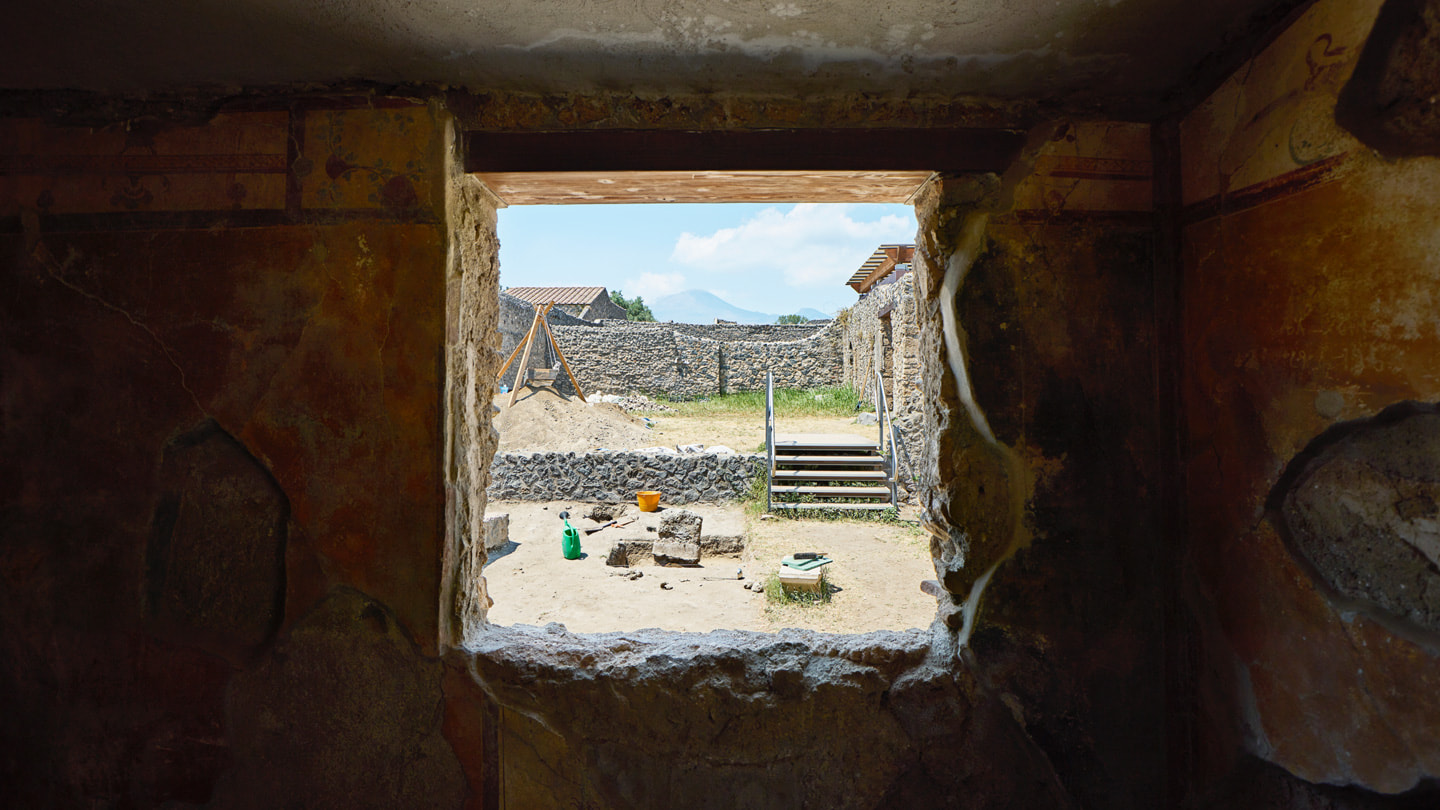 The view through a window at the excavation site shows walls made of stones and Mount Vesuvius in the distance. 
