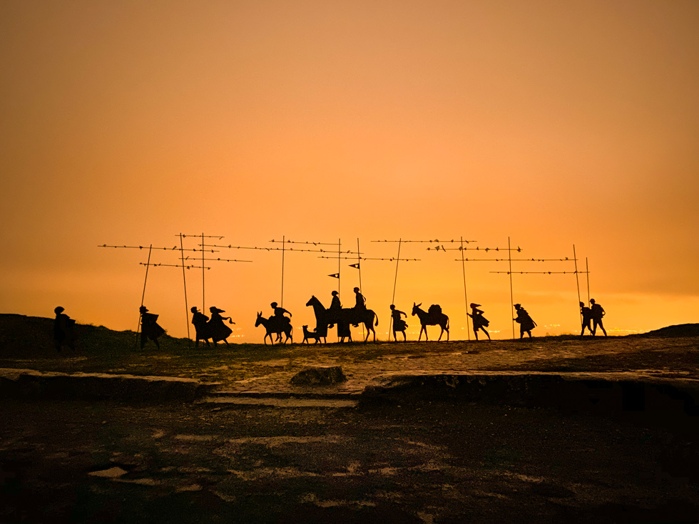 Silhouetten van pelgrims die tegen een oranje gloed door een rotslandschap wandelen.