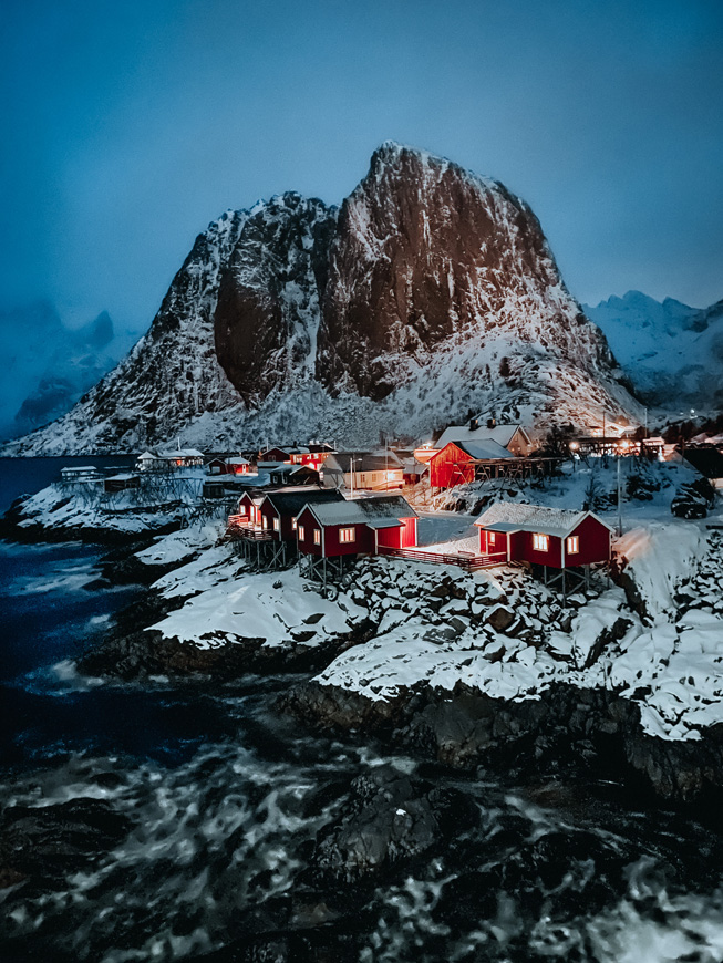 En liten landsby med røde småhus ved havet, med et snødekt fjell i bakgrunnen