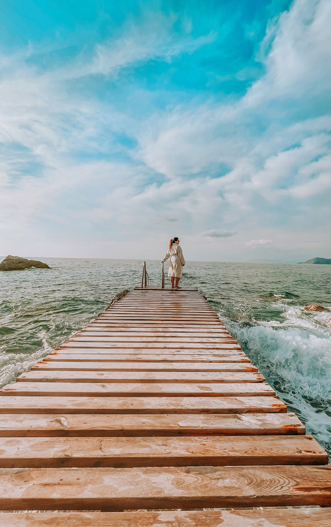 A woman standing at end of a dock facing water.