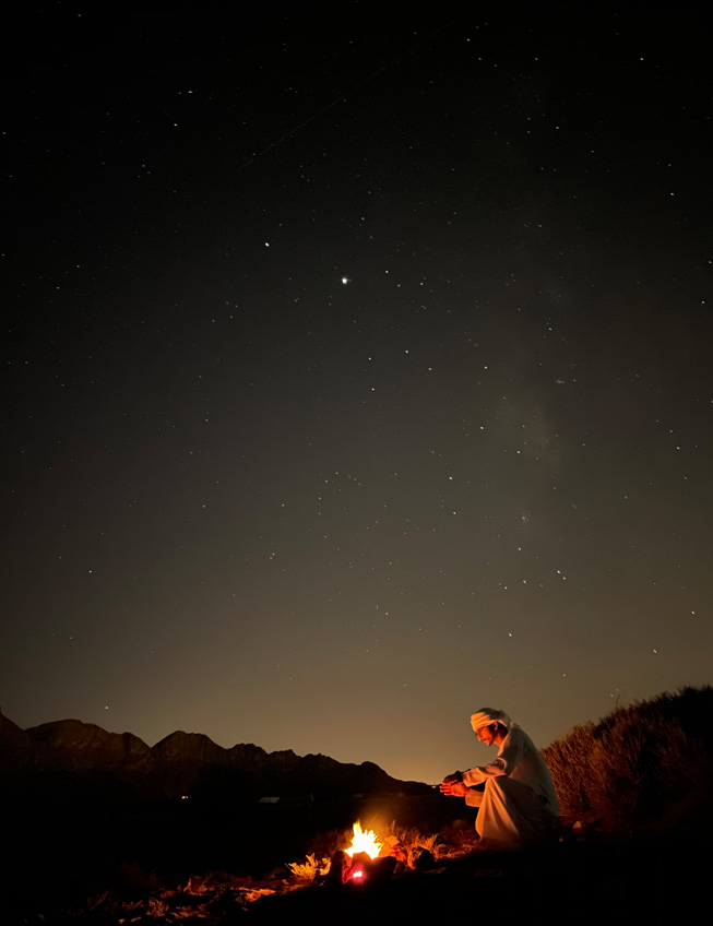 Una toma nocturna de un hombre sentado frente a una fogata en el desierto.