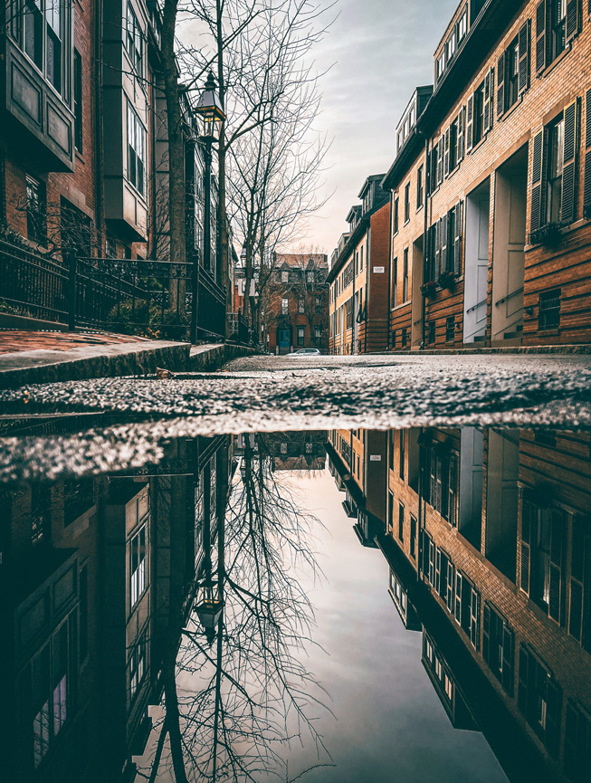 A large puddle of water on the street reflects its surroundings and the sky above.