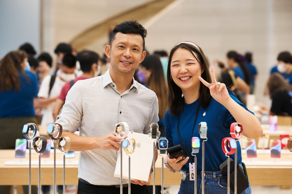 Ein Kunde in Apple Orchard Road posiert mit seinem Einkauf und einem Teammitglied hinter Apple Watch Geräten.
