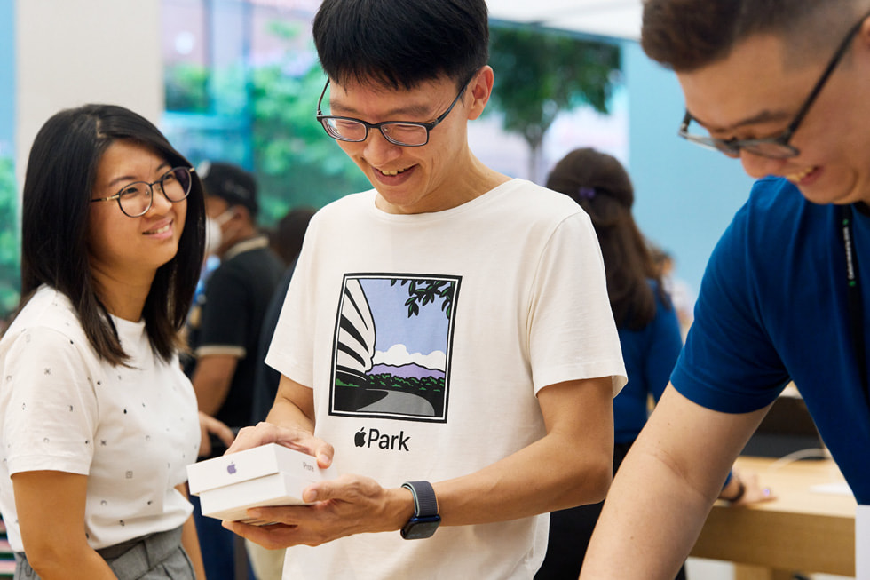 En Apple Orchard Road-kund öppnar förpackningen med en iPhone 14 medan en vän tittar på och en medarbetare ler.