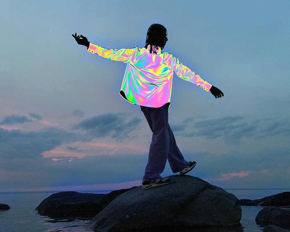 Una foto scattata con iPhone che ritrae una persona con indosso una maglietta metallizzata che si appoggia a una roccia nell’oceano.