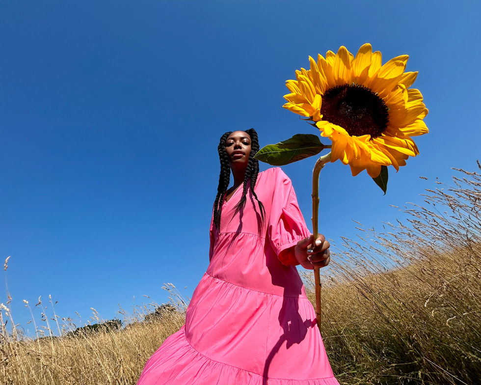 A person in a pink dress holds a sunflower up to the camera in this image shot on iPhone.