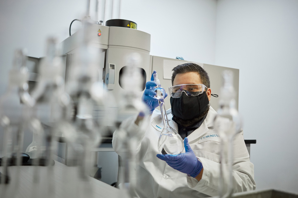 A Corning technician, outfitted in goggles, mask, and gloves, handles glass beakers.