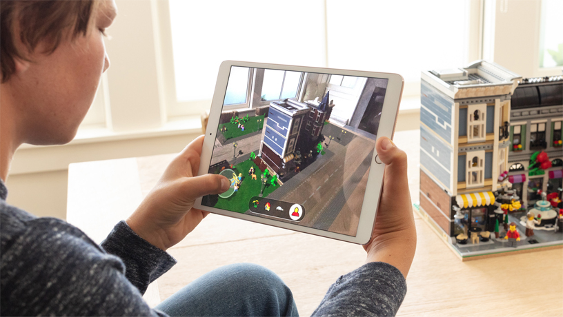 LEGO AR City가 실행된 iPad를 들고, 방 안에서 LEGO 건물 앞에 앉아있는 소년
