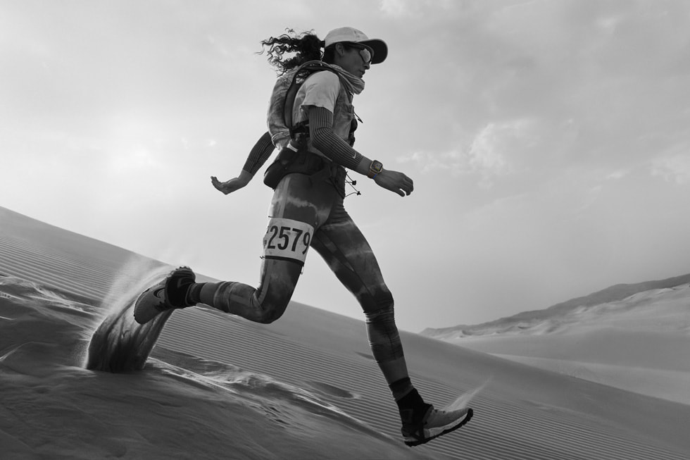An athlete wearing Apple Watch Ultra and a race number runs downhill in sand.