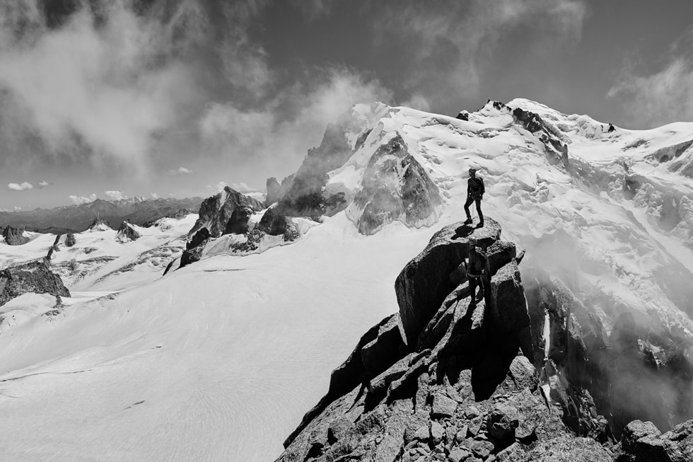 An Apple Watch Ultra user stands on a rugged peak, surrounded by snow-covered mountains. 