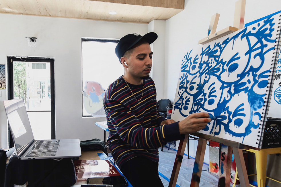 An artist works on an abstract painting in his studio with MacBook open next to him.