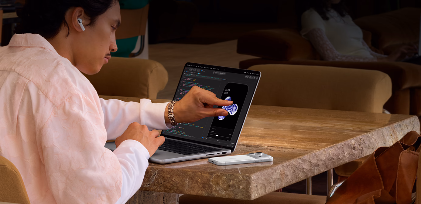 บุคคลหนึ่งนั่งอยู่ที่โต๊ะในที่สาธารณะ โดยใช้คำสั่งหนีบนิ้วเพื่อปรับองค์ประกอบใน Xcode โดยใช้คุณสมบัติการสัมผัสของ MacBook Pro สีเงินพร้อมจอภาพ PROLED XDR, iPhone อยู่บนโต๊ะข้างๆ แล็ปท็อป และบุคคลดังกล่าวกำลังใช้ AirPods Pro