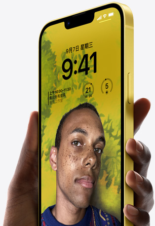 一隻手握著黃色 iPhone 14，手機上的個人化鎖定畫面顯示人物在黃色背景前的照片，並搭配時間與小工具