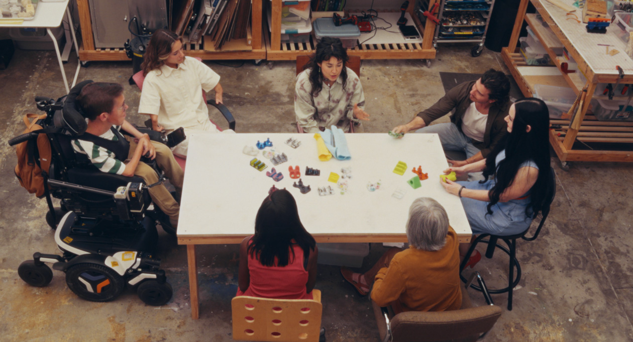 A group of people with different mobility needs sitting together at a table while looking at an array of colourful adaptive smartphone accessories