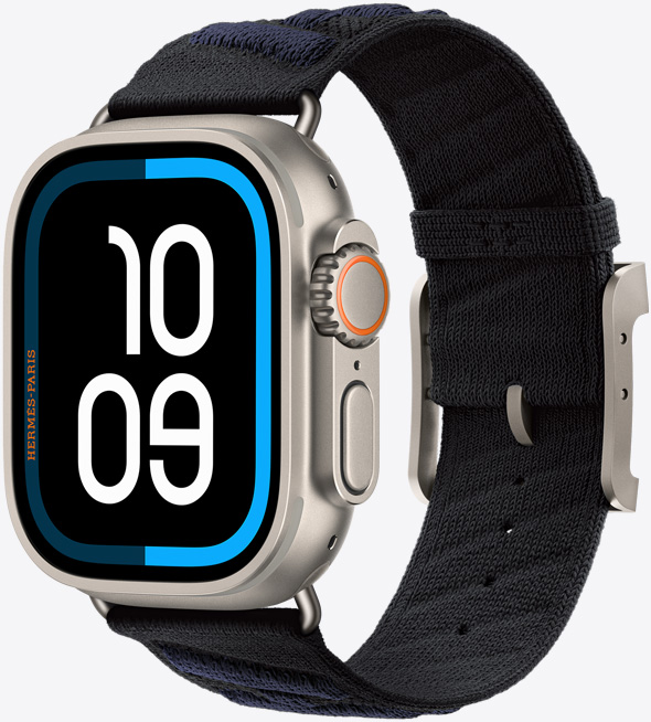 Apple Watch Hermès Ultra 3, caja de titanio, color natural, pantalla de borde a borde, esquinas redondeadas, lateral derecho, Digital Crown, micrófono, botón lateral elevado, esfera digital (10:09), con la correa En Mer, color Noir/Bleu Nuit (negro con franjas diagonales en relieve de color azul oscuro), confeccionada con hilo de poliamida tejido, orificios de ajuste en el centro, hebilla y componentes de titanio en color natural