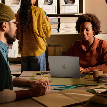 Colleagues collaborating together at a work desk. A cutting mat and other design tools are on the work desk. One colleague interacts with a MacBook.