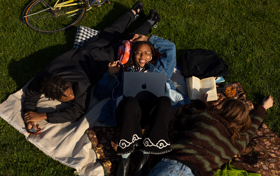 Tiga mahasiswa bersantai di taman. Seorang mahasiswa memakai iPhone. Seorang mahasiswa memakai MacBook Air dan headphone Apple. Seorang mahasiswa memegang buku bersampul tipis.