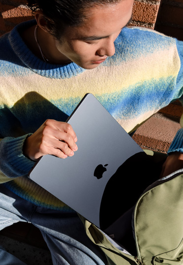 Ein Student, der draußen sitzt. Er holt ein geschlossenes MacBook aus seinem Rucksack.