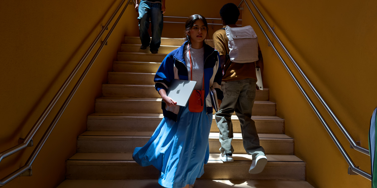 Una studentessa che scende le scale. Tiene in mano un MacBook e alcuni libri.