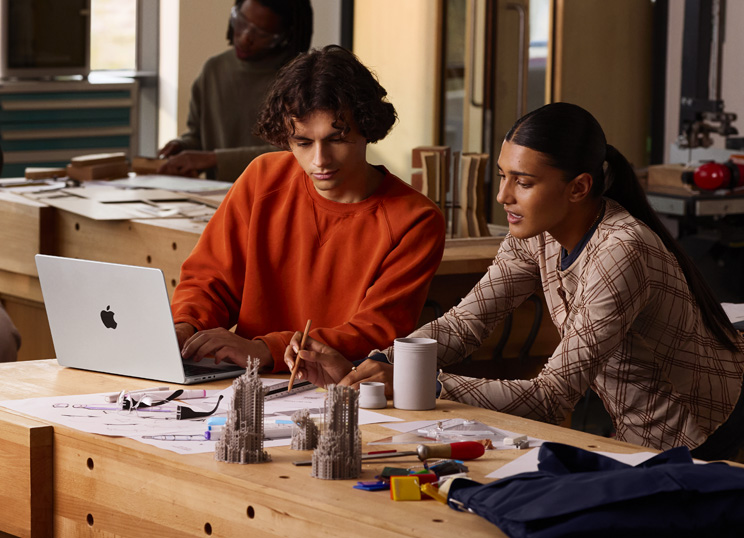 Dos personas colaboran en un proyecto en una mesa, una escribe en un MacBook, la otra usa un lápiz y una regla sobre un papel, hay materiales de arte alrededor.