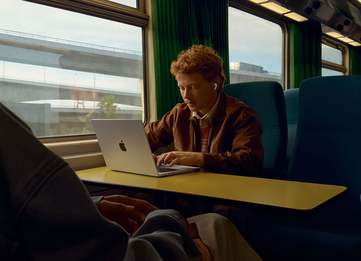 Una persona en el asiento de un tren junto a la ventana usa unos AirPods y trabaja en un MacBook.