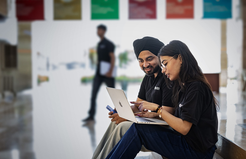 Two students sit in a university hallway while working on a MacBook.