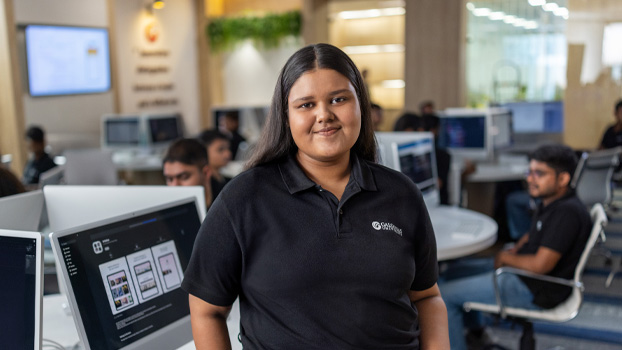 Sharon Bruce stands at a classroom table, smiling at the camera. Behind her, an iMac shows the App Store page for her app.