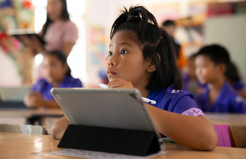 A young girl sits at her desk, looking toward the front of the classroom and fully engaged. She is using an iPad and holding an Apple Pencil.