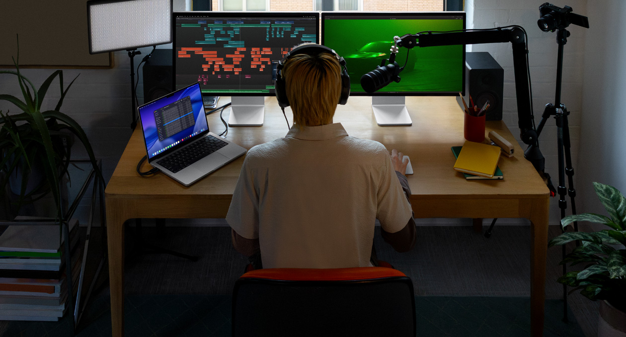 Pessoa sentada à mesa com microfone e fones de ouvido e três aparelhos à sua frente: um monitor Mac Studio Display com ferramentas de edição de áudio, clipes de som em diferentes blocos de cor, um segundo monitor Mac Studio Display com a imagem de um carro na neblina com iluminação verde, a tela do MacBook Pro de 16 polegadas mostrando o app Finder na Mesa, com arquivos do Final Cut Pro, Dock, fundo de mesa elegante azul
