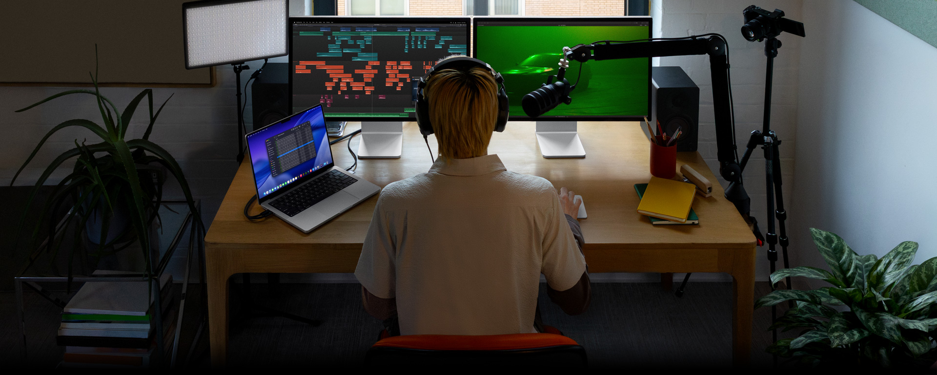Pessoa sentada à mesa com microfone e fones de ouvido e três aparelhos à sua frente: um Mac Studio Display com ferramentas detalhadas de edição de áudio, clipes de som em diferentes blocos de cor, um segundo Mac Studio Display com um carro em iluminação verde, a tela do MacBook Pro de 16 polegadas com uma longa lista de tomadas em um projeto do Final Cut Pro