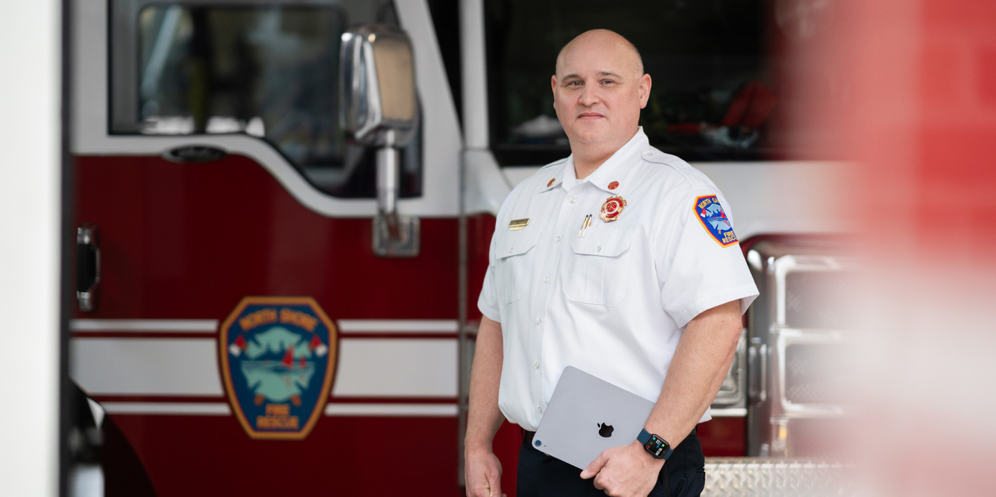 The fire chief stands in front of a fire truck holding an iPad and wearing an Apple Watch