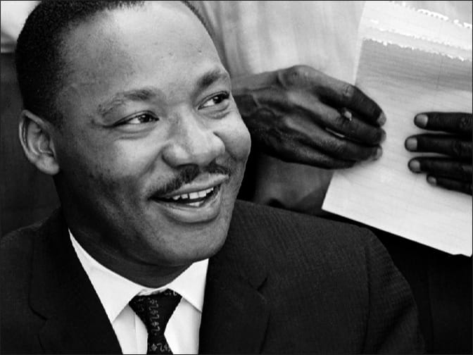 Black and white photo of Dr. Martin Luther King Jr. holding a news conference in Birmingham, Alabama, May 1963, smiling, hands behind him, holding paper with handwritten notes.