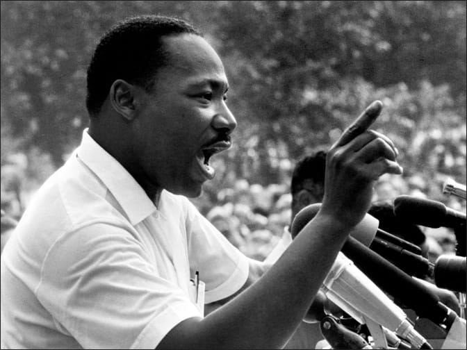 Black and white photo of Dr. Martin Luther King Jr. speaking into microphones outdoors at the March Against Fear rally in Jackson, Mississippi, June 1966, crowd listens.