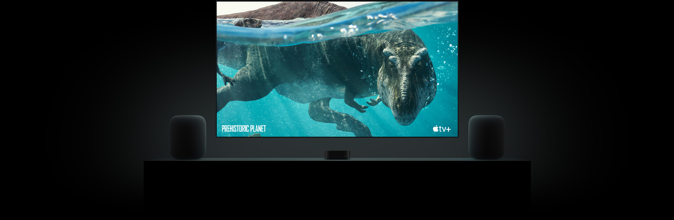 TV màn hình phẳng lớn có hình ảnh sống động của một con khủng long từ Prehistoric Planet.  TV được treo phía trên Apple TV và được đóng khung bởi hai loa HomePod đặt trên bảng điều khiển phòng khách