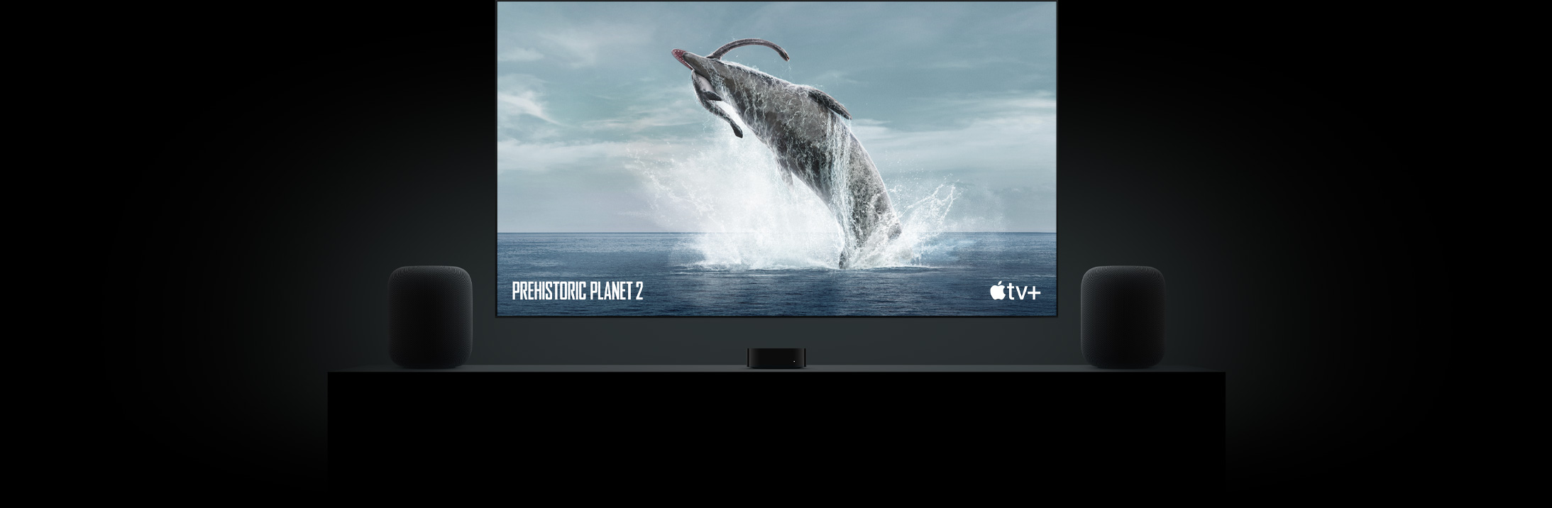 Большой телевизор с плоским экраном с ярким изображением динозавра с доисторической планеты.  Телевизор подвешен над Apple TV и обрамлен двумя динамиками HomePod, расположенными на консоли в гостиной.