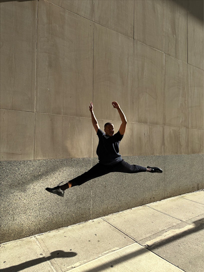 Photo d’un homme faisant un saut en grand écart devant un mur de béton dans des conditions de faible éclairage. Le sujet se découpe en contre-jour, mais le mur et le sol sont lumineux et détaillés.