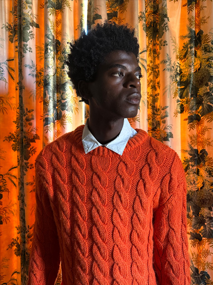 Een foto van een man in een felrode trui die voor een gordijn met een patroon staat. De foto is bij weinig licht gemaakt met de hoofdcamera.