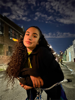 Ar nakts režīmu uzņemta bilde, kurā sieviete pozē ar divriteni pilsētas ielā naktī.