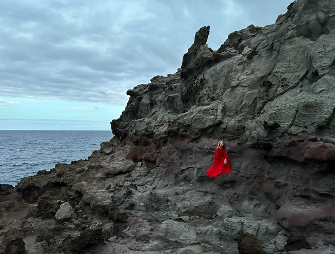 빨간색 드레스를 입고 바닷가 바위를 배경으로 포즈를 취하고 있는 여성을 메인 카메라로 촬영한 저조도 사진.