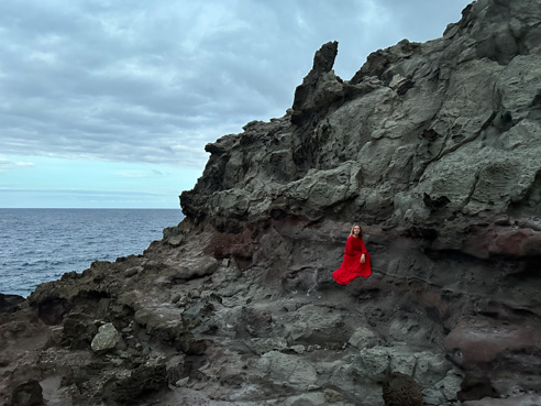 Ein beeindruckendes Foto von einer Person in Rot, die sich von einer grauen Felsküste abhebt, aufgenommen mit der Pro Level Hauptkamera.