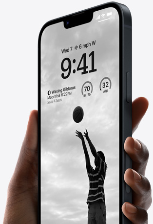 A hand holding iPhone 14 in Midnight with a personalized Lock Screen featuring a photo of someone playing basketball, black font, and widgets visible