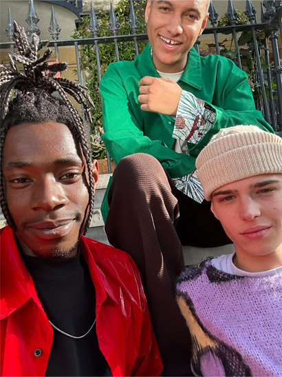 Ein Selfie mit drei Personen, die in farbenfrohen, miteinander kontrastierenden Outfits auf Stufen sitzen, aufgenommen mit der TrueDepth Kamera.