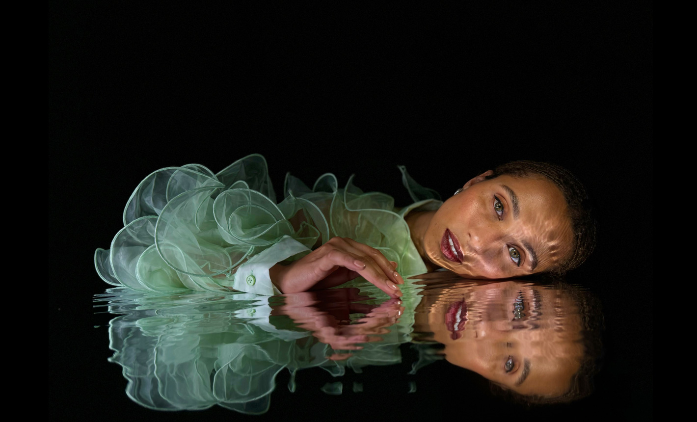 A beautifully detailed photo shot on iPhone 15 Pro of a woman laying close to her reflection in rippled water on a black background