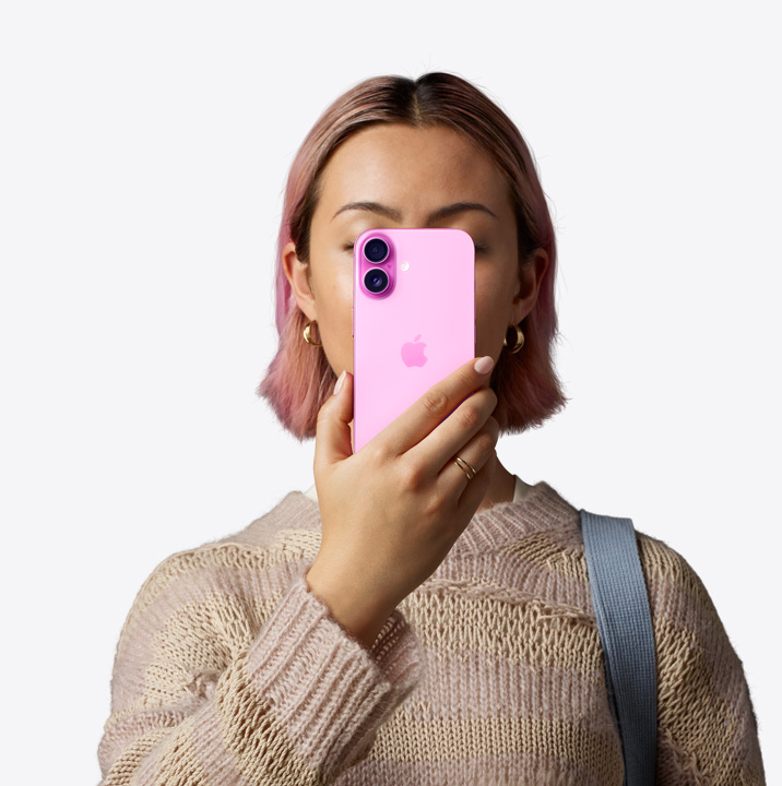 Egy ember rózsaszín iPhone 16-ot tart az arca elé, mutatva a készülék hátoldalát a bal felső sarkában lévő duplakamera-rendszerrel.