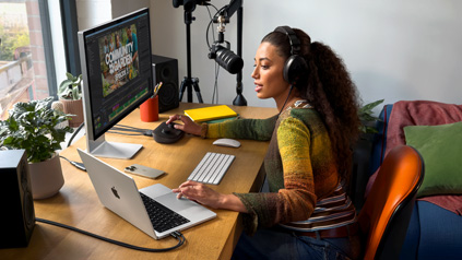 Een vrouw in haar woning achter haar bureau met daarop een MacBook Pro verbonden met een extern display. Ze draagt een koptelefoon en spreekt in een microfoon.