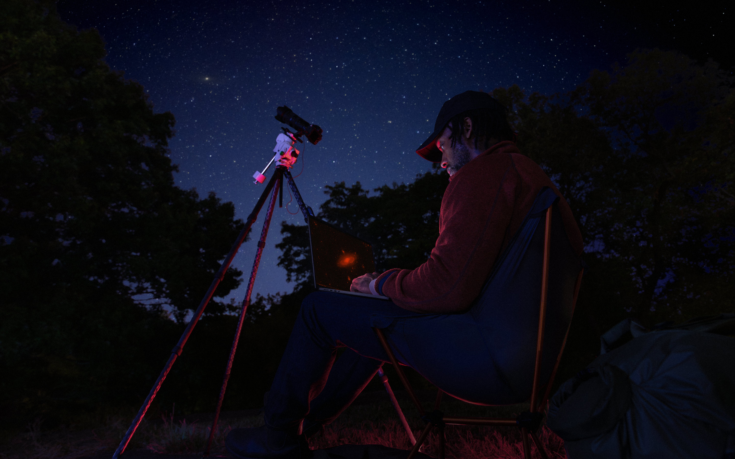 A person outside at night using a MacBook Pro on their lap, connected to a device on a tripod pointed toward the starry sky