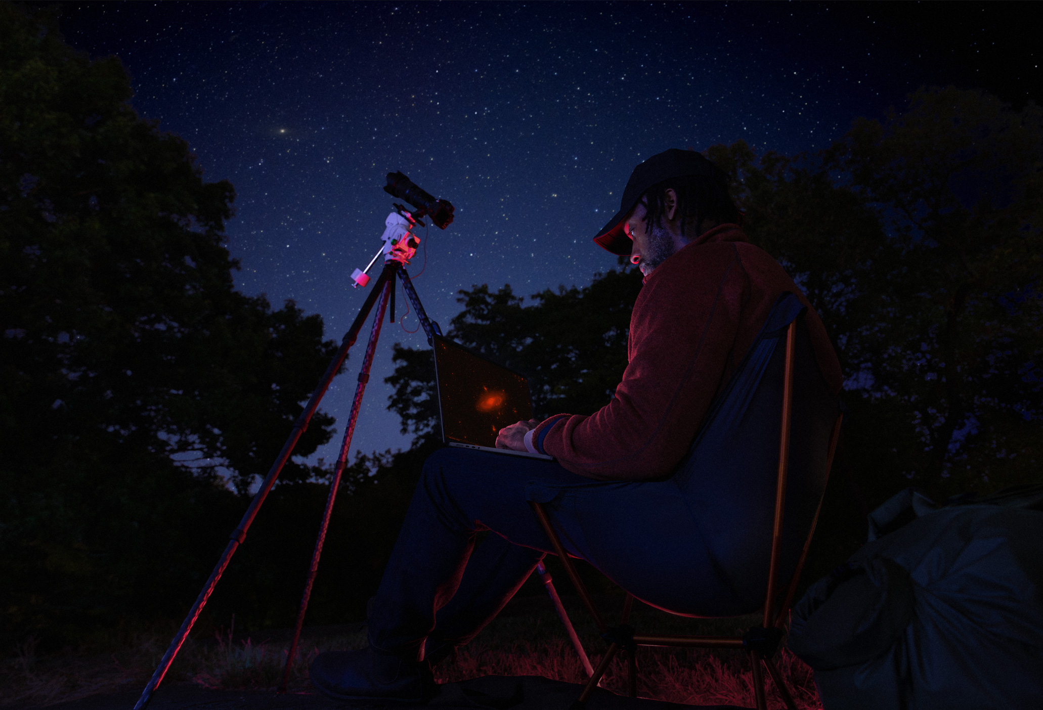 夜間戶外，一個人將 MacBook Pro 放在腿上使用，筆電連接三腳架上對著星空的裝置