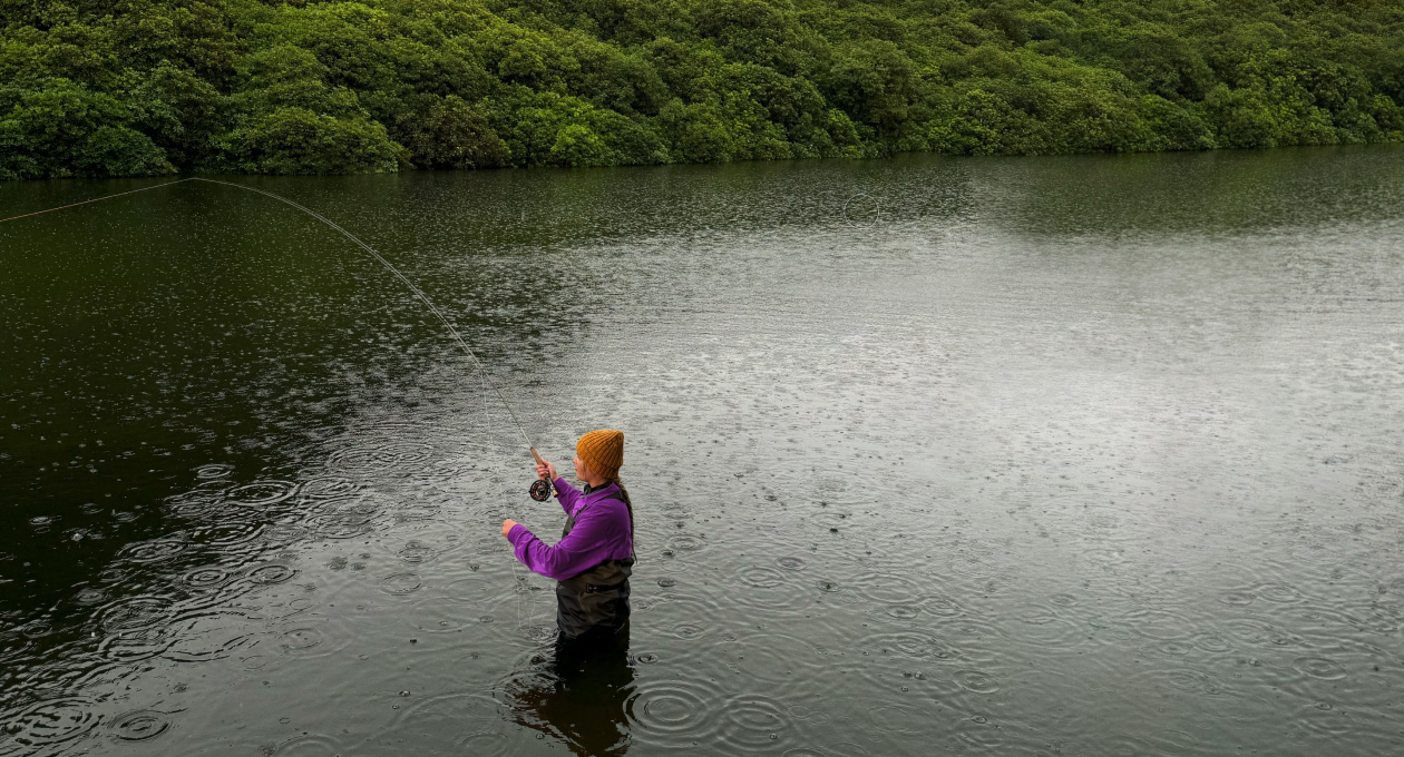 Foto seorang perempuan sedang melakukan fly-fishing di sebuah danau, alat Pixelmator Pro menghilangkan objek tidak diinginkan di latar belakang yang disorot dengan warna kuning